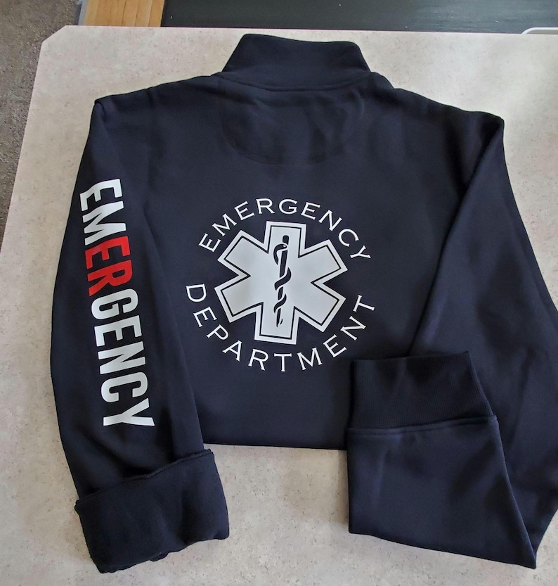 May include: Black zippered jacket with long sleeves and white text on the sleeves and back. The text on the sleeves reads "EMERGENCY". The text on the back reads "EMERGENCY DEPARTMENT" with a white star of life graphic.