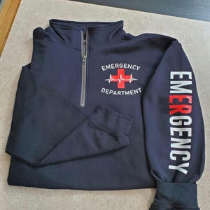 May include: Navy blue quarter-zip sweatshirt with "EMERGENCY DEPARTMENT" printed on the front and "EMERGENCY" printed on the sleeve. The design includes a red cross and a heartbeat line. The sweatshirt has a silver zipper.