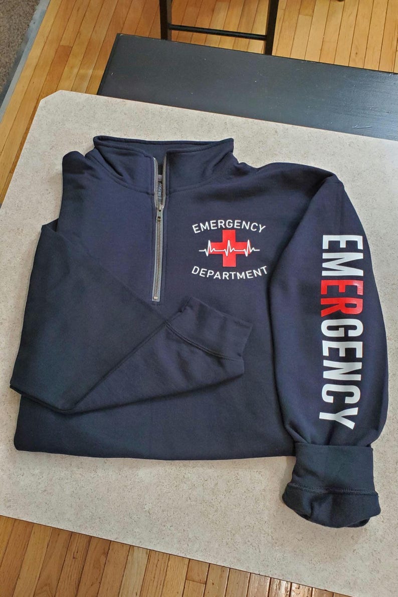 May include: Navy blue quarter-zip sweatshirt with "EMERGENCY DEPARTMENT" printed on the front and "EMERGENCY" printed on the sleeve. The design includes a red cross and a heartbeat line. The sweatshirt has a silver zipper.