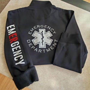 May include: Black zippered sweatshirt with the words "Emergency Department" printed on the back and the word "Emergency" printed on the left sleeve. The back features a white star of life symbol with a snake and staff.