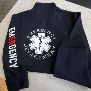 May include: Black zippered jacket with a white star of life logo and the text "Emergency Department" on the back. The left sleeve has the word "Emergency" printed vertically in white with a red stripe.