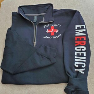 May include: Navy blue quarter-zip sweatshirt with "EMERGENCY DEPARTMENT" printed on the front and "EMERGENCY" down the sleeve. The design includes a red cross and heartbeat graphic. The sweatshirt has a gray collar.