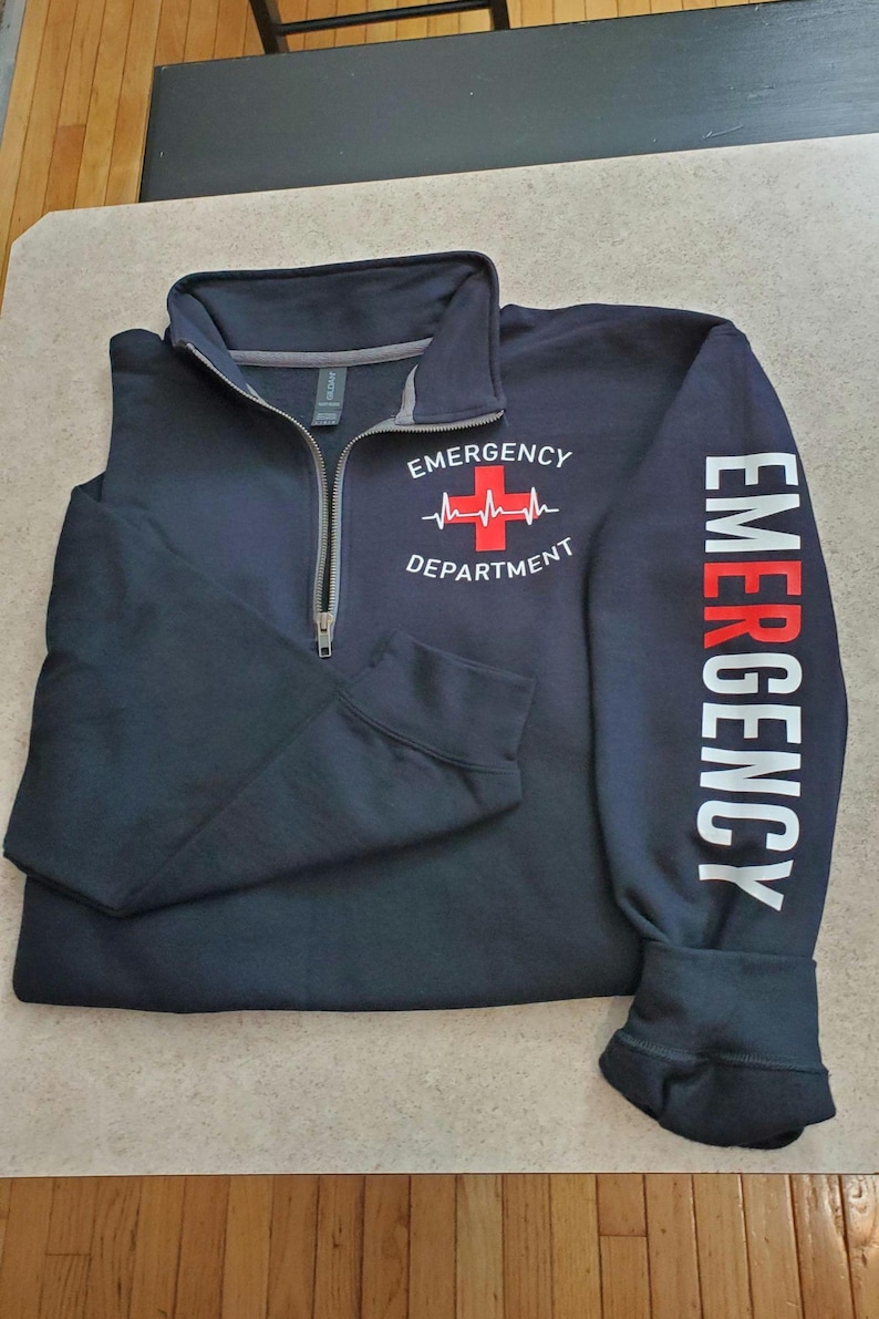May include: Navy blue quarter-zip sweatshirt with "EMERGENCY DEPARTMENT" printed on the front and "EMERGENCY" down the sleeve. The design includes a red cross and heartbeat graphic. The sweatshirt has a gray collar.