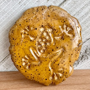 May include: A ceramic wall hanging in the shape of a frog. The frog is brown with a speckled glaze and has a raised design.