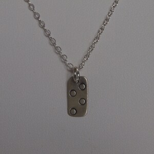 Puede incluir: Un collar plateado con un colgante rectangular. El colgante presenta cuatro pequeños círculos espaciados uniformemente. La cadena es delicada y el collar se muestra sobre un fondo neutro.