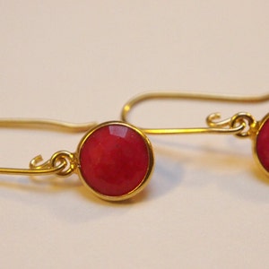 Op de afbeelding: Een paar goudkleurige oorhangers met ronde, facetgeslepen, rode edelstenen. De oorbellen hebben een eenvoudig, elegant ontwerp met een haaksluiting. De edelstenen zijn gezet in een gouden bezel.