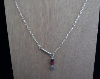 May include: A silver necklace with a delicate chain and a horizontal bar pendant. Below the bar hangs a cluster of small red and black beads, and a small flower charm. The necklace is displayed against a light brown wooden surface.