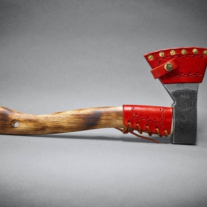 May include: A handcrafted axe with a wooden handle and a metal blade. The axe head is protected by a red leather sheath, and the handle is wrapped with a red leather grip. The axe is designed for chopping wood and other outdoor tasks.