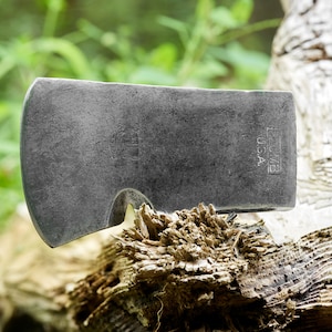 May include: A close-up of a gray metal axe head with a small hole in the side. The axe head is resting on a piece of weathered wood. The text "Estwing USA" is stamped on the axe head.