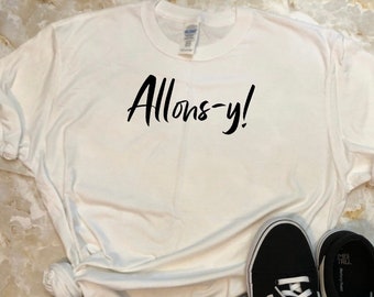 Puede incluir: Camiseta blanca con el texto "Allons-y!" impreso en negro. La camiseta se lleva con jeans azules y zapatillas negras.