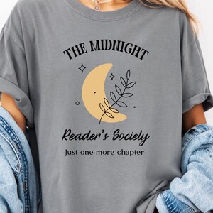 May include: Gray t-shirt with the text "The Midnight Reader's Society" and a crescent moon graphic. The shirt also includes the phrase "Just one more chapter". The shirt is a comfortable, casual style.