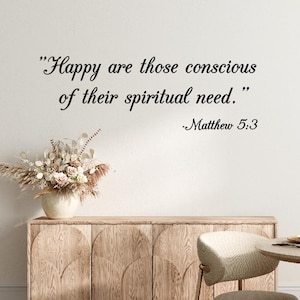 May include: A room with a light-colored wooden cabinet, a round table, and two chairs. The wall features the quote "Happy are those conscious of their spiritual need." from Matthew 5:3. A vase of dried flowers sits on the cabinet.