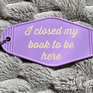 May include: Purple motel key chain with a silver key ring. The keychain has the text "I closed my book to be here" in yellow.