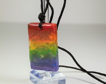 Puede incluir: Un colgante de colores arcoíris con un cordón negro. El colgante está hecho de resina y tiene un efecto de capas, con colores rojo, naranja, amarillo, verde, azul y morado. El colgante está en un expositor transparente.