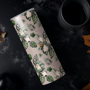 K&ouml;nnte beinhalten: Ein Edelstahlbecher mit einem skurrilen Halloween-Design. Der Becher zeigt ein Muster aus wei&szlig;en Geistern, gr&uuml;nen Bl&auml;ttern und einem kleinen orangefarbenen K&uuml;rbis auf hellbeigem Hintergrund. Der Becher ist zylindrisch und hat einen silbernen Deckel.