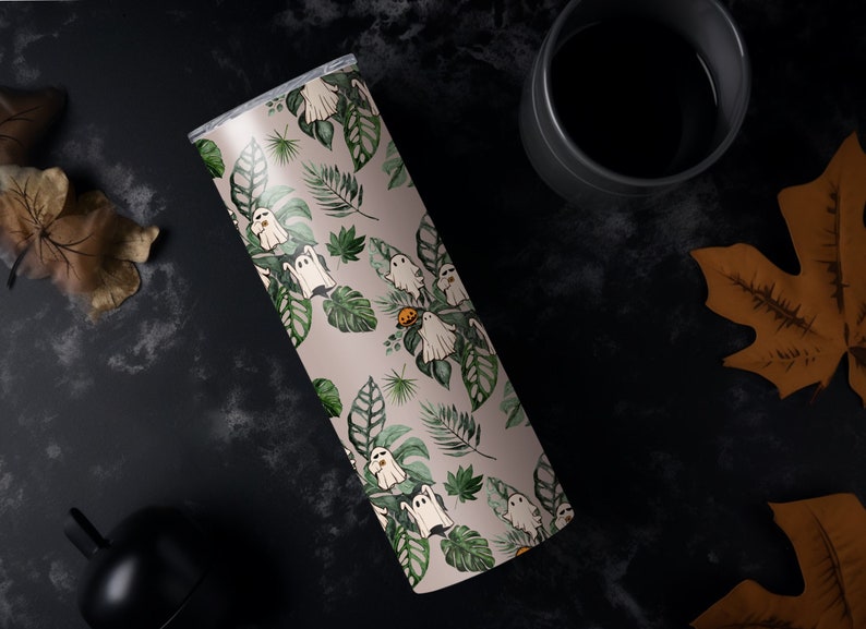 K&ouml;nnte beinhalten: Ein Edelstahlbecher mit einem skurrilen Halloween-Design. Der Becher zeigt ein Muster aus wei&szlig;en Geistern, gr&uuml;nen Bl&auml;ttern und einem kleinen orangefarbenen K&uuml;rbis auf hellbeigem Hintergrund. Der Becher ist zylindrisch und hat einen silbernen Deckel.