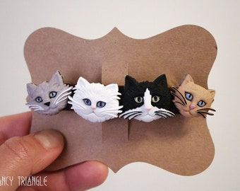 May include: A hair clip featuring four sculpted cat heads in gray, white, black and white, and tan colors, each with detailed whiskers and eyes. The cat heads are attached to a brown cardboard display card with the text THE FANCY TRIANGLE printed at the bottom.