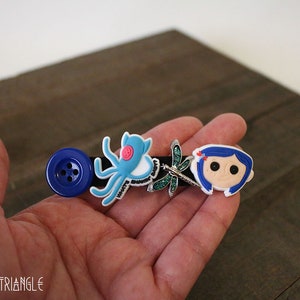 Puede incluir: Una horquilla negra con un bot&oacute;n azul grande, un pulpo azul claro, una lib&eacute;lula y un personaje de dibujos animados. La horquilla se sostiene en una mano sobre un fondo de madera.