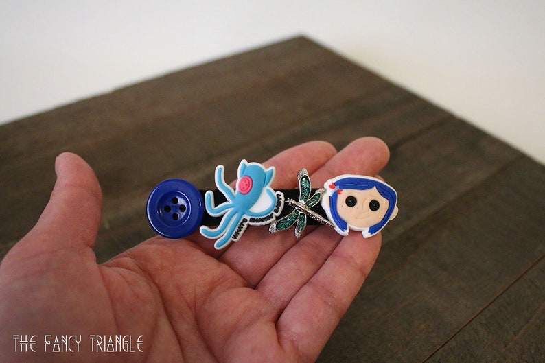 Puede incluir: Una horquilla negra con un bot&oacute;n azul grande, un pulpo azul claro, una lib&eacute;lula y un personaje de dibujos animados. La horquilla se sostiene en una mano sobre un fondo de madera.