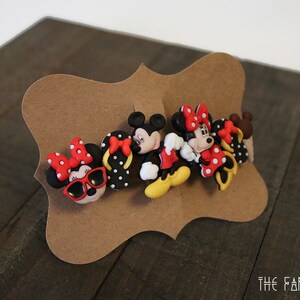 May include: A set of six colorful Minnie Mouse and Mickey Mouse character buttons. The buttons are made of plastic and feature a variety of poses and expressions. The buttons are displayed on a brown card with the text "THE FANCY TRIANGLE" printed on the bottom.