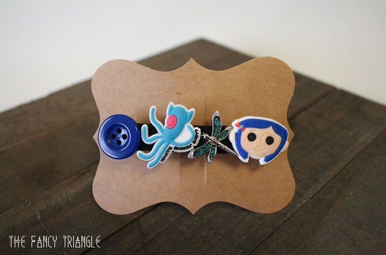 Puede incluir: Una pinza para el pelo negra con un bot&oacute;n azul, un pulpo azul, una lib&eacute;lula y una cara de personaje de dibujos animados. La pinza se muestra en una tarjeta marr&oacute;n. El texto "THE FANCY TRIANGLE" est&aacute; en la parte inferior.
