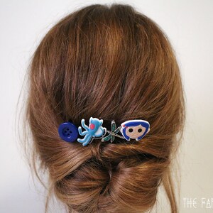 Puede incluir: Una horquilla decorativa para el cabello con un bot&oacute;n azul, un pulpo azul, una lib&eacute;lula y un personaje de dibujos animados. La horquilla est&aacute; colocada sobre cabello casta&ntilde;o peinado en un mo&ntilde;o.
