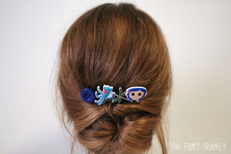 Puede incluir: Una horquilla decorativa para el cabello con un bot&oacute;n azul, un pulpo azul, una lib&eacute;lula y un personaje de dibujos animados. La horquilla est&aacute; colocada sobre cabello casta&ntilde;o peinado en un mo&ntilde;o.