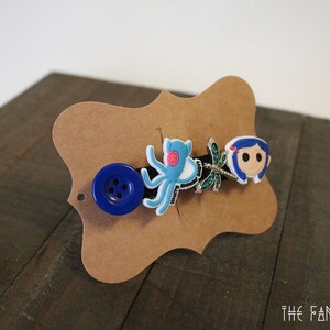 Puede incluir: Una pinza para el pelo decorativa con un bot&oacute;n azul, un pulpo azul claro, una lib&eacute;lula y un personaje de dibujos animados con pelo azul. La pinza est&aacute; montada en una tarjeta marr&oacute;n con forma decorativa.