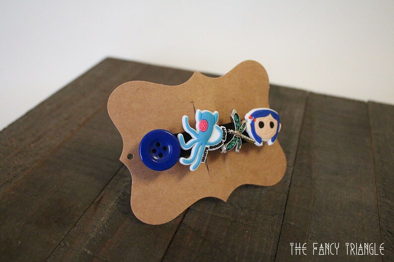 Puede incluir: Una pinza para el pelo decorativa con un bot&oacute;n azul, un pulpo azul claro, una lib&eacute;lula y un personaje de dibujos animados con pelo azul. La pinza est&aacute; montada en una tarjeta marr&oacute;n con forma decorativa.
