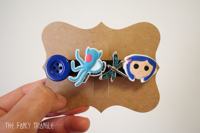 Puede incluir: Una pinza de pelo negra con un bot&oacute;n azul, un pulpo azul claro, una lib&eacute;lula y la cara de una ni&ntilde;a de dibujos animados. El pulpo tiene el texto "WHAT'S SHAKIN' BABY?". La pinza est&aacute; en una tarjeta marr&oacute;n.