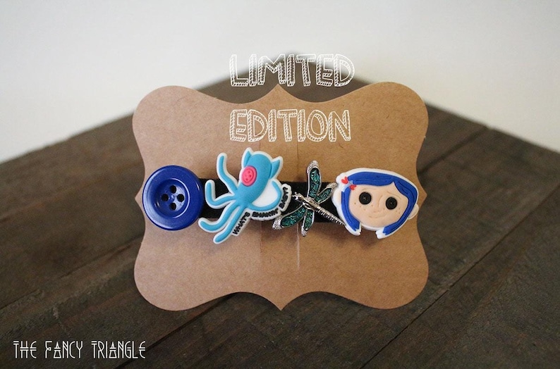 Puede incluir: Una pinza para el pelo negra con un bot&oacute;n azul grande, un pulpo azul claro, una lib&eacute;lula y una cara de dibujos animados con pelo azul. Las palabras "LIMITED EDITION" est&aacute;n encima de la pinza en una tarjeta marr&oacute;n.