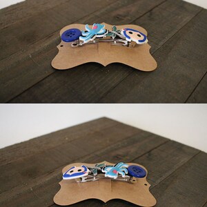 Puede incluir: Dos horquillas decorativas sobre una tarjeta marr&oacute;n. Una presenta un bot&oacute;n azul, una flor azul y un personaje de dibujos animados. La otra tiene un pulpo azul y un personaje de dibujos animados. Las horquillas son plateadas.