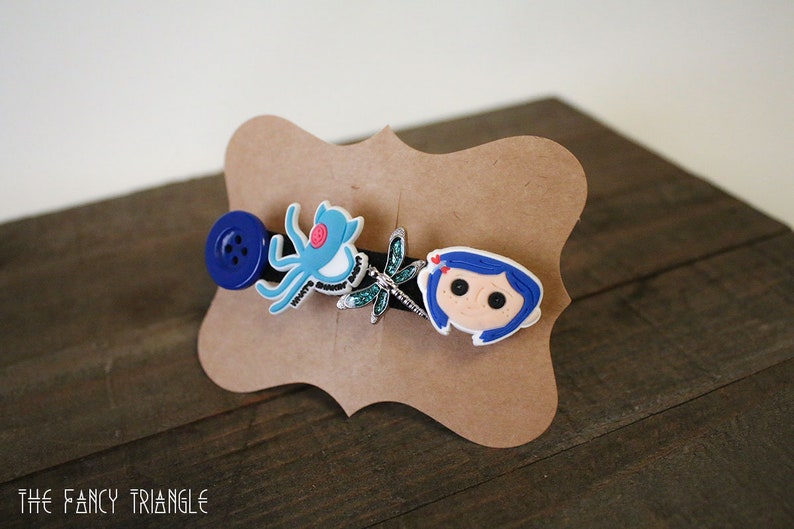 Puede incluir: Una horquilla decorativa para el cabello con un bot&oacute;n azul, un pulpo azul, una lib&eacute;lula y un personaje de dibujos animados. La horquilla est&aacute; montada sobre una tarjeta marr&oacute;n y expuesta sobre una superficie de madera.