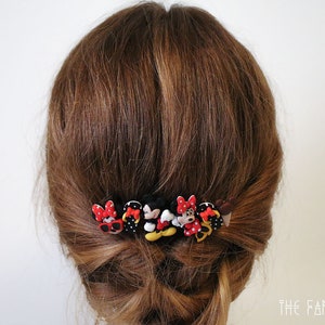 May include: A set of six hair clips featuring cartoon characters with red, black, and yellow accents. The clips are arranged in a row and are attached to a person's brown hair.