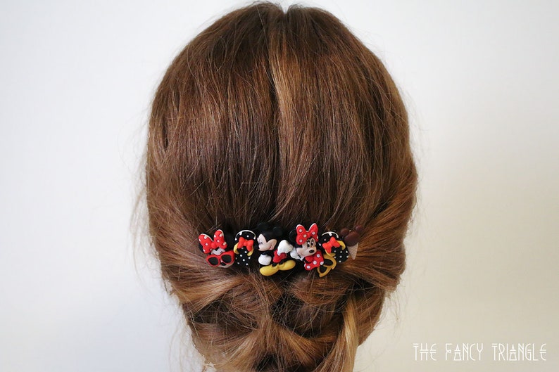 May include: A set of six hair clips featuring cartoon characters with red, black, and yellow accents. The clips are arranged in a row and are attached to a person's brown hair.