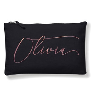 Peut inclure: Pochette noire rectangulaire avec le nom "Olivia" en écriture rose doré. La pochette a une fermeture éclair sur le dessus. Le texte est centré sur la pochette.