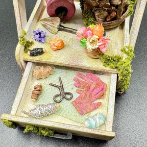 May include: A miniature wooden garden cart filled with gardening tools and floral arrangements. The cart features a small trowel, scissors, pink gloves, and a variety of colorful flowers. The cart is decorated with faux moss.