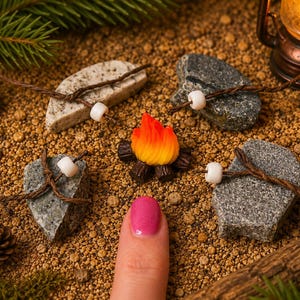 Puede incluir: Una figura de fogata en miniatura con llamas naranjas y amarillas y troncos de color marrón oscuro, rodeada por cuatro piedras grises, cada una envuelta con hilo marrón y una cuenta blanca, dispuestas en círculo sobre una superficie arenosa.