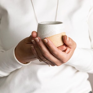 Op de afbeelding: Een kleine, handgemaakte keramische beker met een gespikkelde witte bovenkant en een bruinachtige onderkant. De beker wordt met beide handen vastgehouden, wat het eenvoudige, moderne ontwerp laat zien. De beker is ongeveer 7.6 cm hoog.