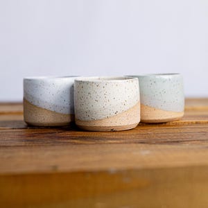 May include: Three handmade ceramic cups with a speckled, light-colored glaze and a natural, unglazed base. The cups are arranged on a wooden surface, showcasing their unique, handcrafted design. The cups have a diagonal color separation.