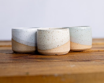 May include: Three handmade ceramic cups with a speckled, light-colored glaze and a natural, unglazed base. The cups are arranged on a wooden surface, showcasing their unique, handcrafted design. The cups have a diagonal color separation.