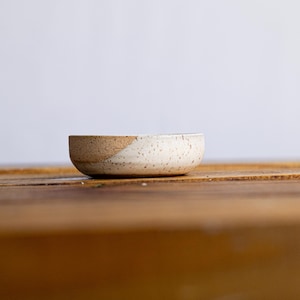 Op de afbeelding: Een kleine, handgemaakte keramische kom met een gespikkelde, gebroken witte glazuur. De rand heeft een diagonale sectie van ongeglazuurde, getextureerde bruine klei. De kom staat op een houten oppervlak, wat het eenvoudige, rustieke ontwerp laat zien.