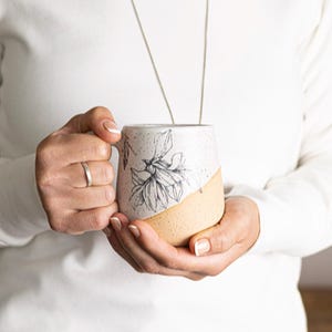 May include: A speckled ceramic mug with a floral design. The mug features a cream-colored upper half with a black floral illustration and a tan-colored lower half. The mug is held by a person wearing a white sweater.