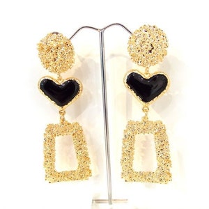 May include: Gold tone dangle earrings featuring a black heart and a square shape. The earrings are textured and have a delicate design.