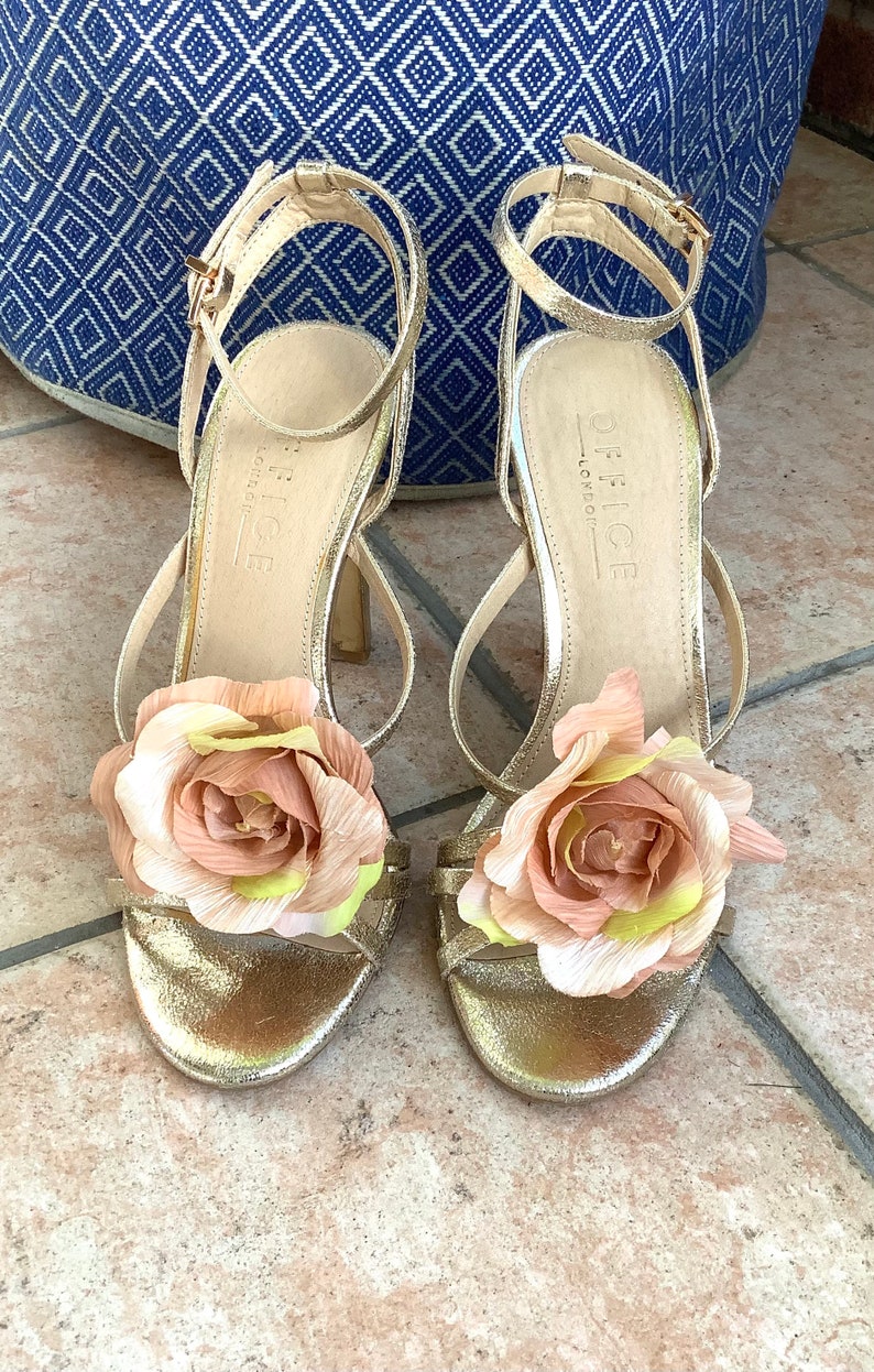 May include: Gold high-heeled sandals with a floral embellishment on each strap. The sandals are made by Office London.