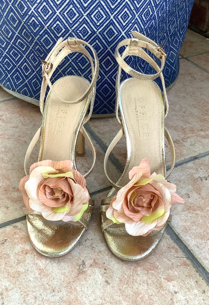 May include: Gold heeled sandals with a large pink flower embellishment on each strap. The sandals are labeled "Office London".