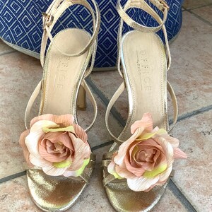 May include: Gold heeled sandals with a large pink flower embellishment on each strap. The sandals are labeled "Office London".