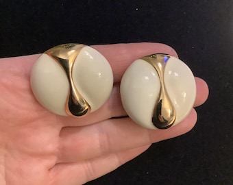 May include: A pair of cream-colored, round earrings with a gold-colored, teardrop-shaped design. The earrings are held in a person's hand against a dark background. The earrings have a glossy finish.