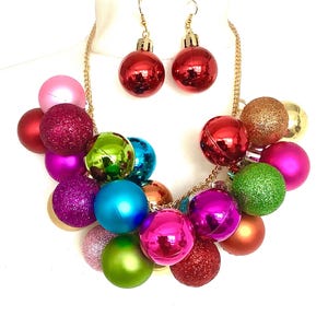 May include: A festive necklace and earring set featuring colorful Christmas ornament-shaped beads. The necklace has a gold chain and a cluster of red, pink, green, and blue ornaments. The earrings are red, round ornaments with gold hooks.