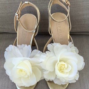 May include: Gold heeled sandals with white flower embellishments. The sandals have ankle straps and the brand name "Office - London" is printed on the insole.